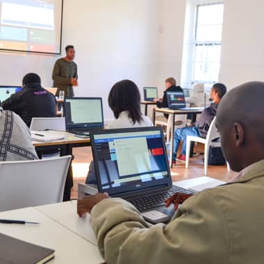 Students working on laptops in a classroom with a facilitator standing in front of the class.